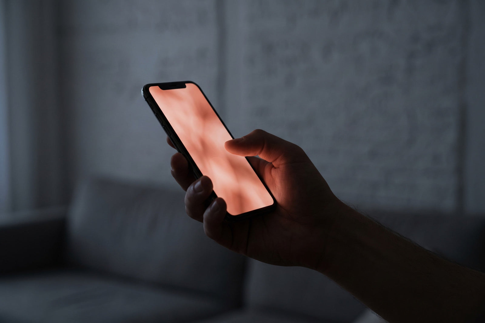 Fotografia editorial em close: uma mão anônima segura um smartphone em ambiente escuro. O brilho da tela ilumina só os dedos e ilumina o vidro em tom terracota suave.